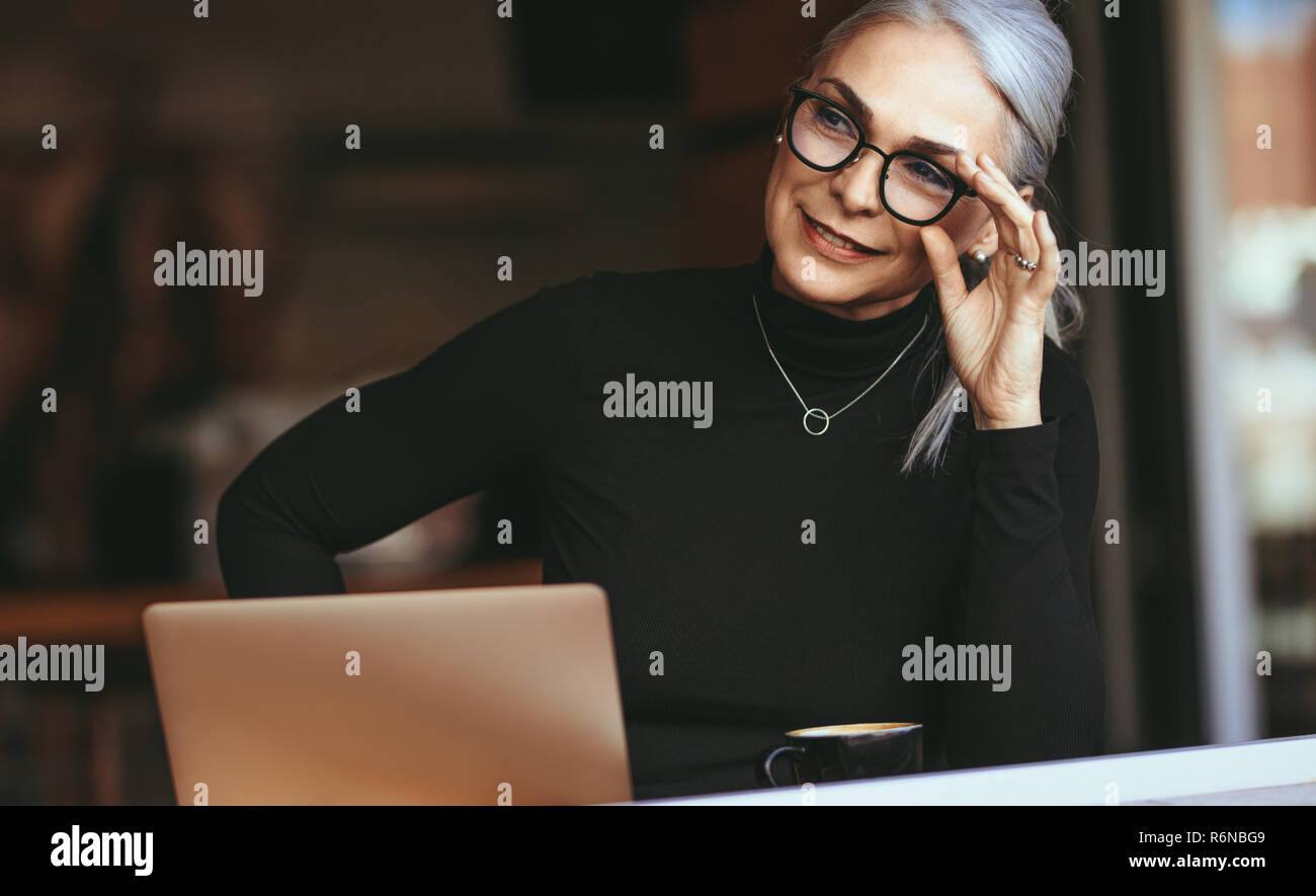 Ritratto di bella senior imprenditrice seduta presso il cafe con il computer portatile che guarda lontano e di pensare. Donna matura presso la caffetteria. Foto Stock