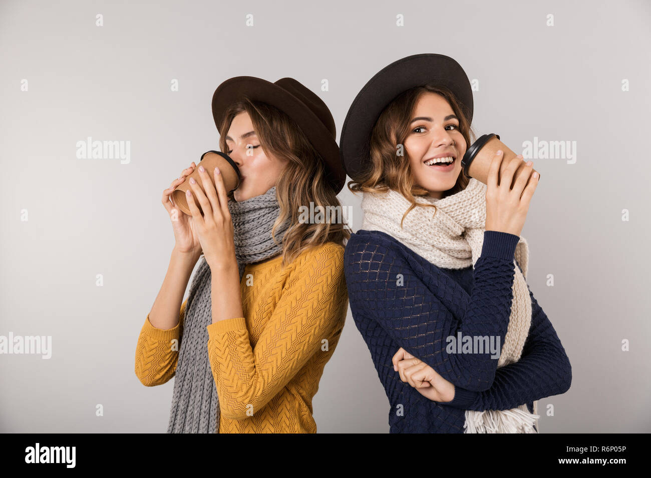 Immagine delle due donne positive indossando cappelli e sciarpe azienda caffè da asporto in bicchieri di carta isolate su uno sfondo grigio Foto Stock