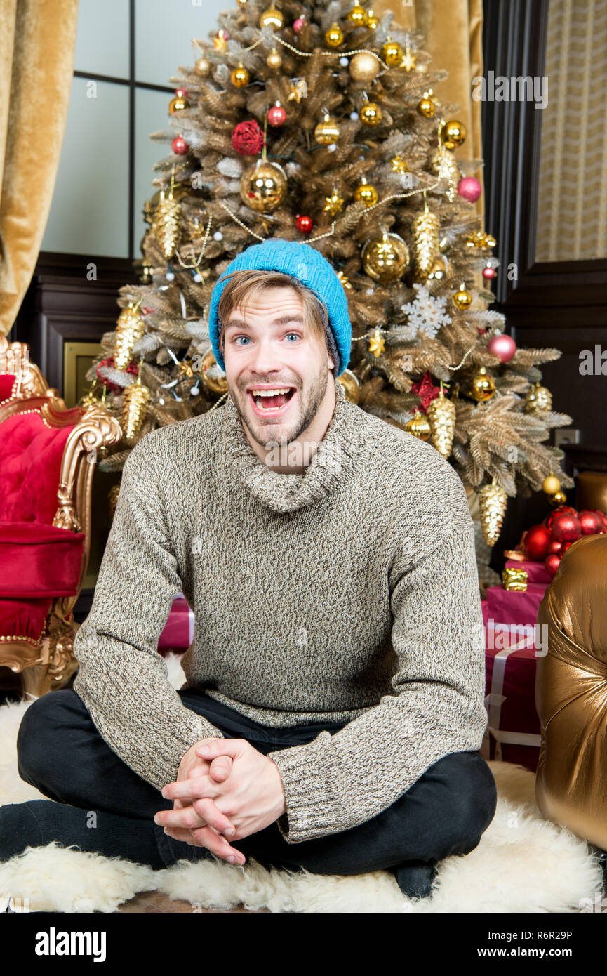 Macho sorriso sul tappeto peloso a albero di natale in camera. Uomo seduto  in un caldo cappello, maglione ad albero di Natale. Vacanze a preparazione  e celebrazione. Felice anno nuovo, eve, partito.