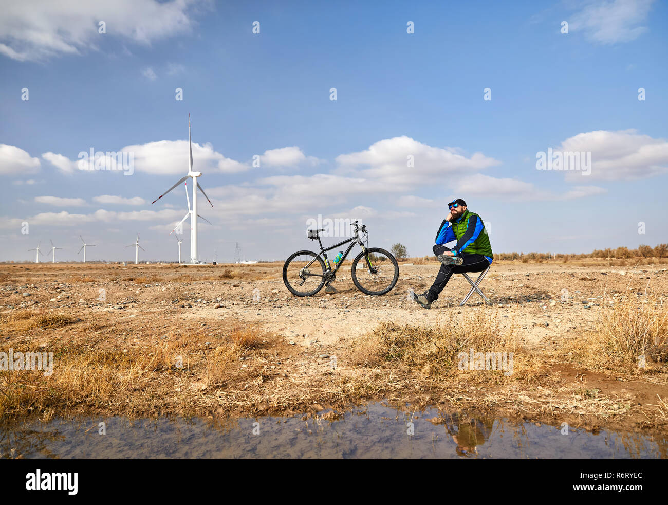 Uomo Barbuto in appoggio sulla sedia con la sua bicicletta di montagna vicino a fattoria eolica nel deserto Foto Stock