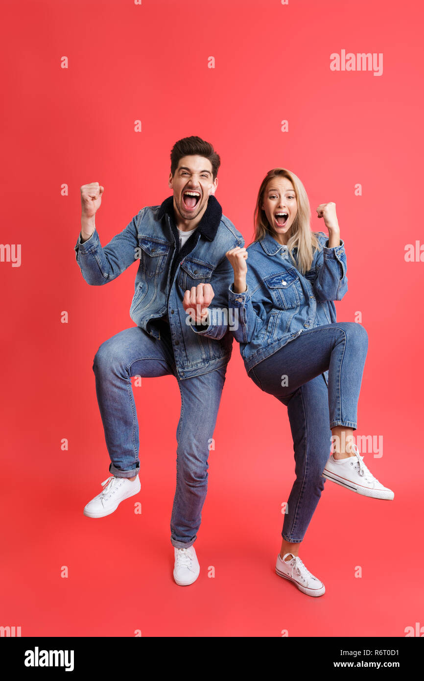 A piena lunghezza Ritratto di una felice coppia giovane vestito in abiti denim isolate su sfondo rosso, celebrando il successo Foto Stock