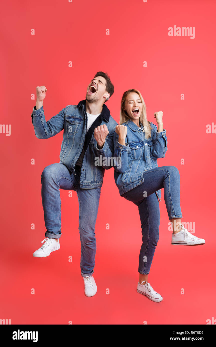 A piena lunghezza Ritratto di un allegro coppia giovane vestito in abiti denim isolate su sfondo rosso, celebrando il successo Foto Stock