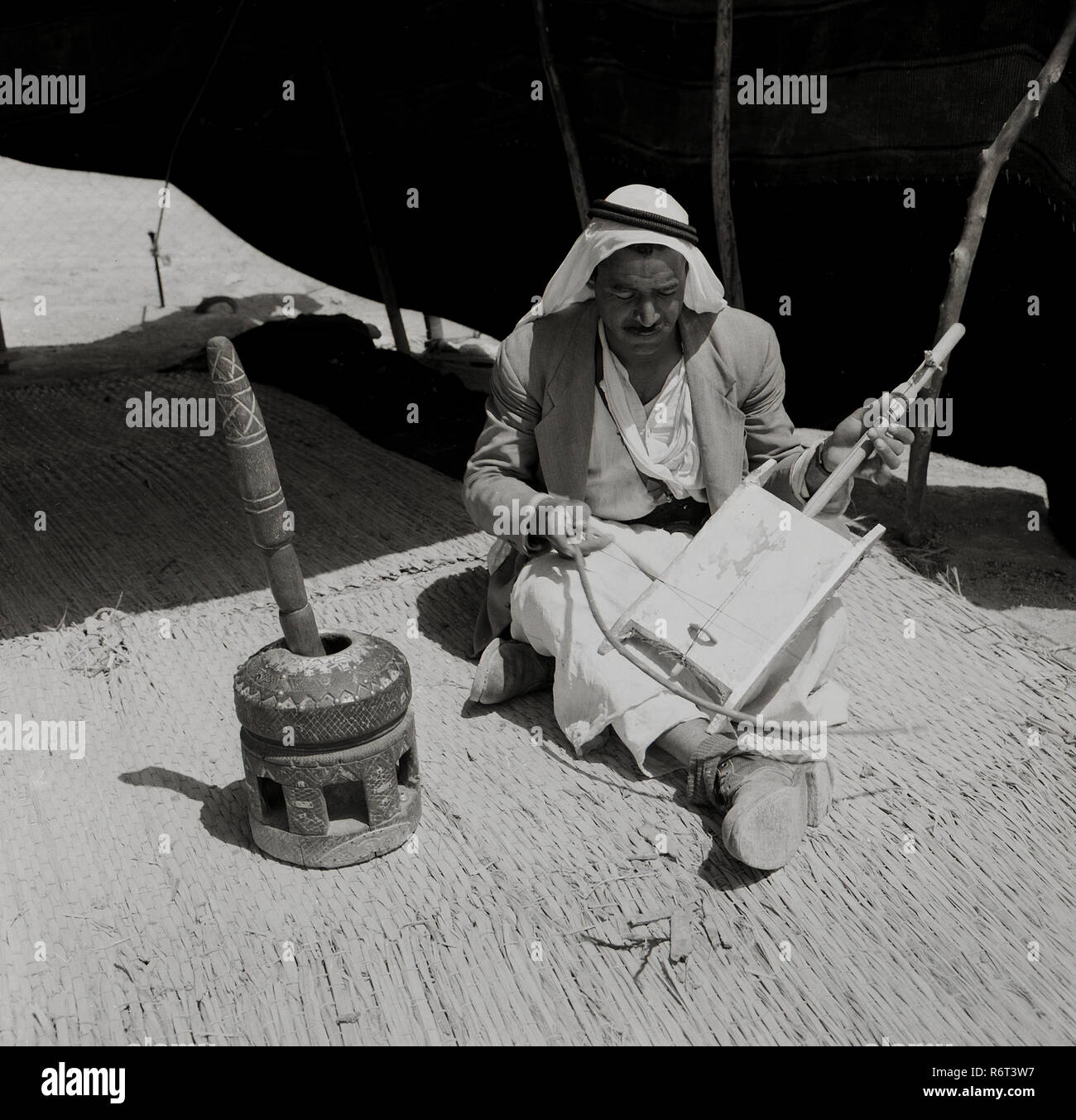 Degli anni Cinquanta, storico, un arabo beduino indossando un keffiyeh headress seduti su un tappeto di reed all'interno della sua tenda a giocare un rebab, un beduino tradizionale strumento musicale, noto come il deserto violino. Il rebab è costituito da un rettangolo di asse di legno ricoperta di pelle - una scatola sonora- e attraverso questo una singola stringa da un cavallo e di coda un crine prua per riprodurlo. Tradizionalmente utilizzato per accompagnare letture di poesie e fortemente connesso con la tribù beduina, la storia e i suoi antenati, il rebab è di grande importanza e molto apprezzato anche nella loro cultura. Foto Stock