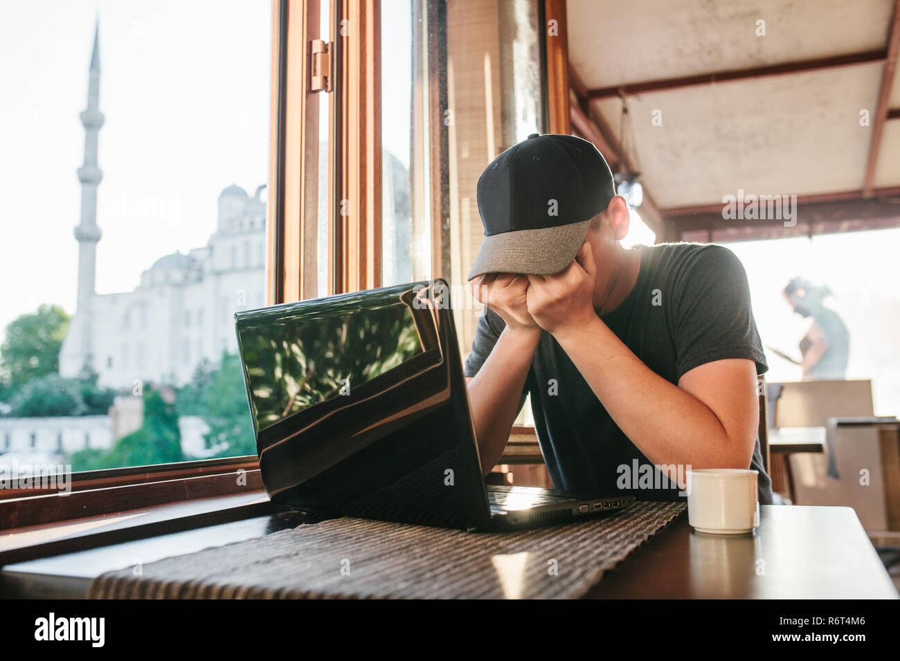 Uomo in cafe si siede a tavola con computer accanto alla finestra e faccia nascosta dalle mani Foto Stock