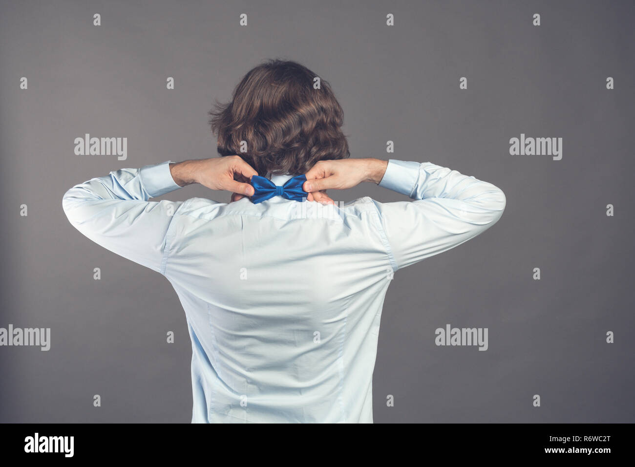 Il sig. perfezione. Vista posteriore del bello shaggy curly giovane uomo che indossa maglietta blu regolando il suo bow tie rimanendo contro uno sfondo grigio. Elegantemente vestito guy. Tonica. Posto per il testo Foto Stock