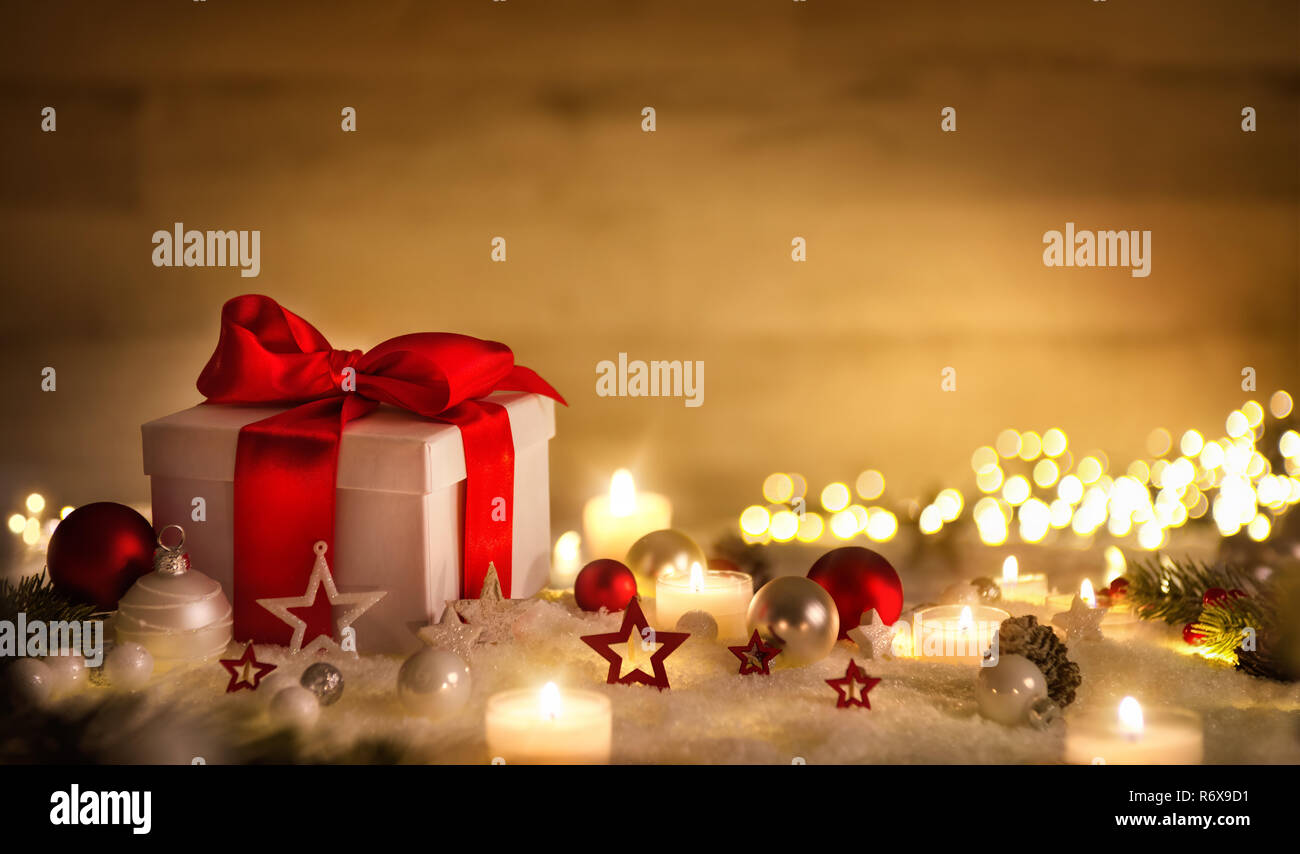 Scena di Natale con una scatola regalo bianca, arco rosso, candele, luci, baubles e neve, marrone con spazio di copia Foto Stock