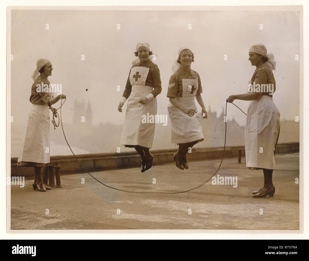 Giovani infermieri volontari della seconda Guerra Mondiale saltano / si rilassano in cima ad un edificio a Londra, Westminster sullo sfondo, indossando un'uniforme volontaria di soccorso (VAD) intorno al 1940, Londra, Regno Unito Foto Stock