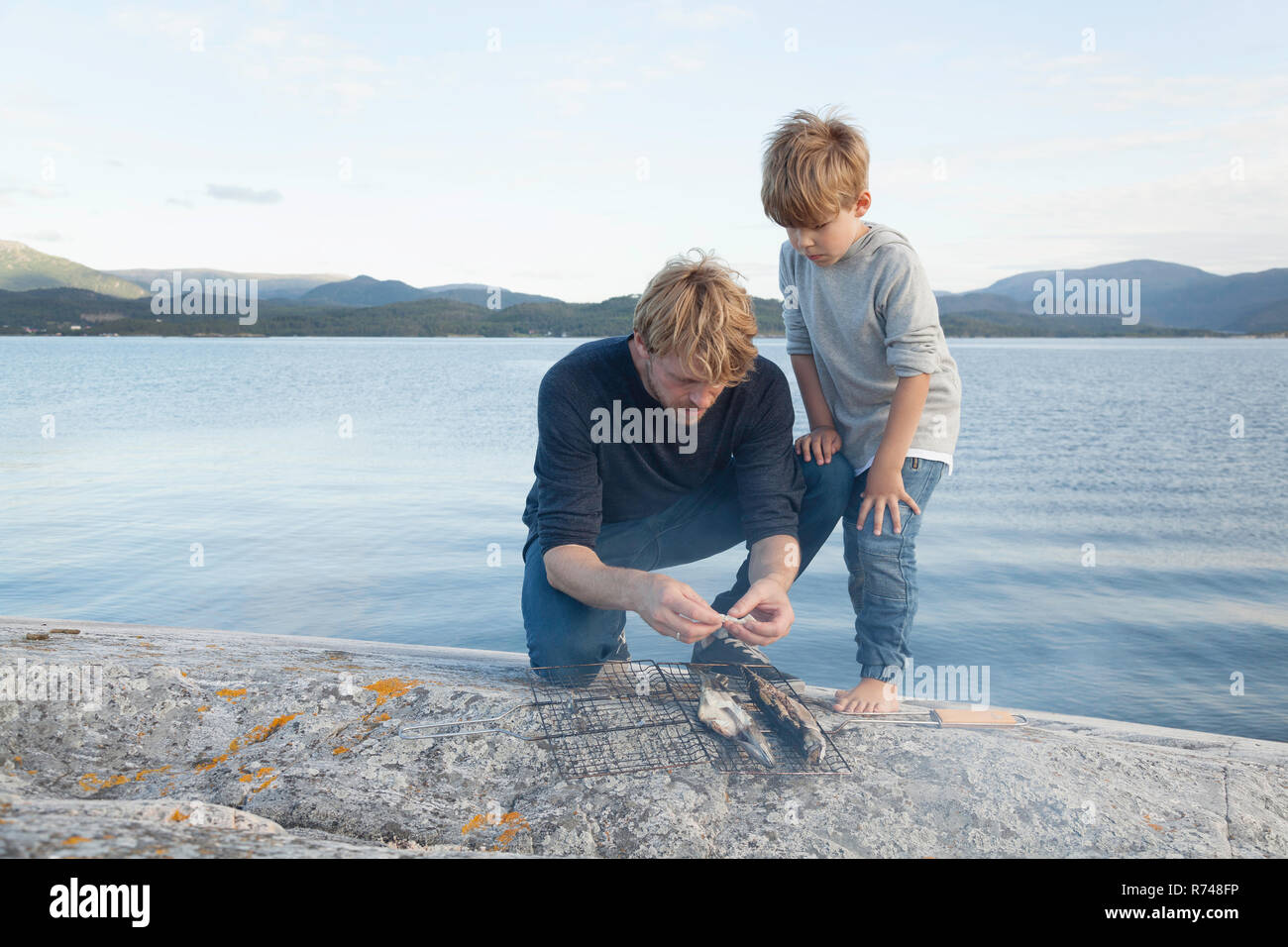 Ragazzo e padre preparare pesce alla griglia su roccia di ingresso, Aure, More og Romsdal, Norvegia Foto Stock