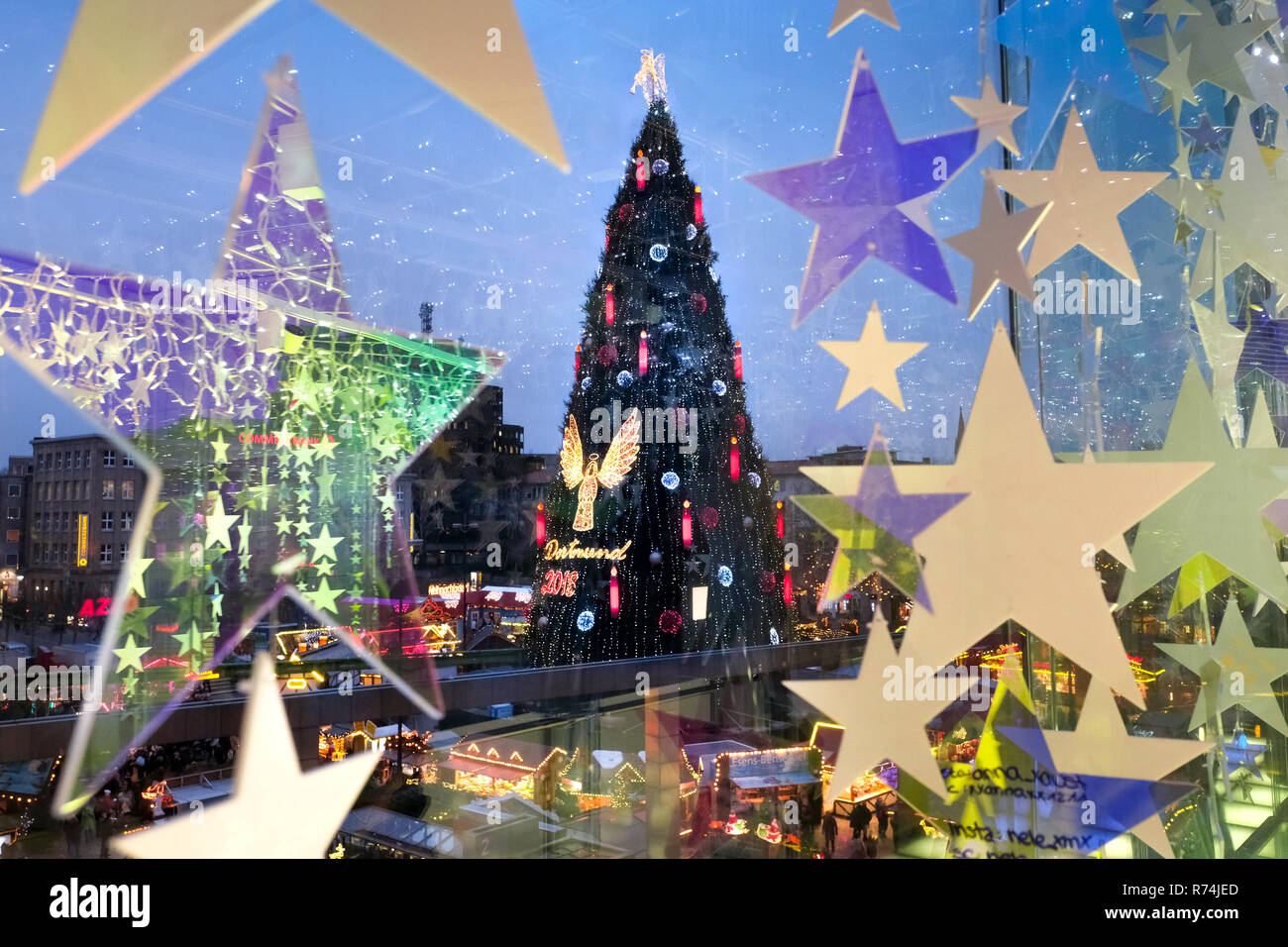 Dortmund, Germania, 07.12.2018 - Vista dell albero di Natale più grande del mondo a Dortmund Mercatino di Natale (45 metri di altezza, fatto di 1.700 abeti rossi, illuminato da 48.000 luci LED e con un angelo in alto) attraverso la finestra di un department store decorate con stelle a Hansaplatz a Dortmund. Foto Stock