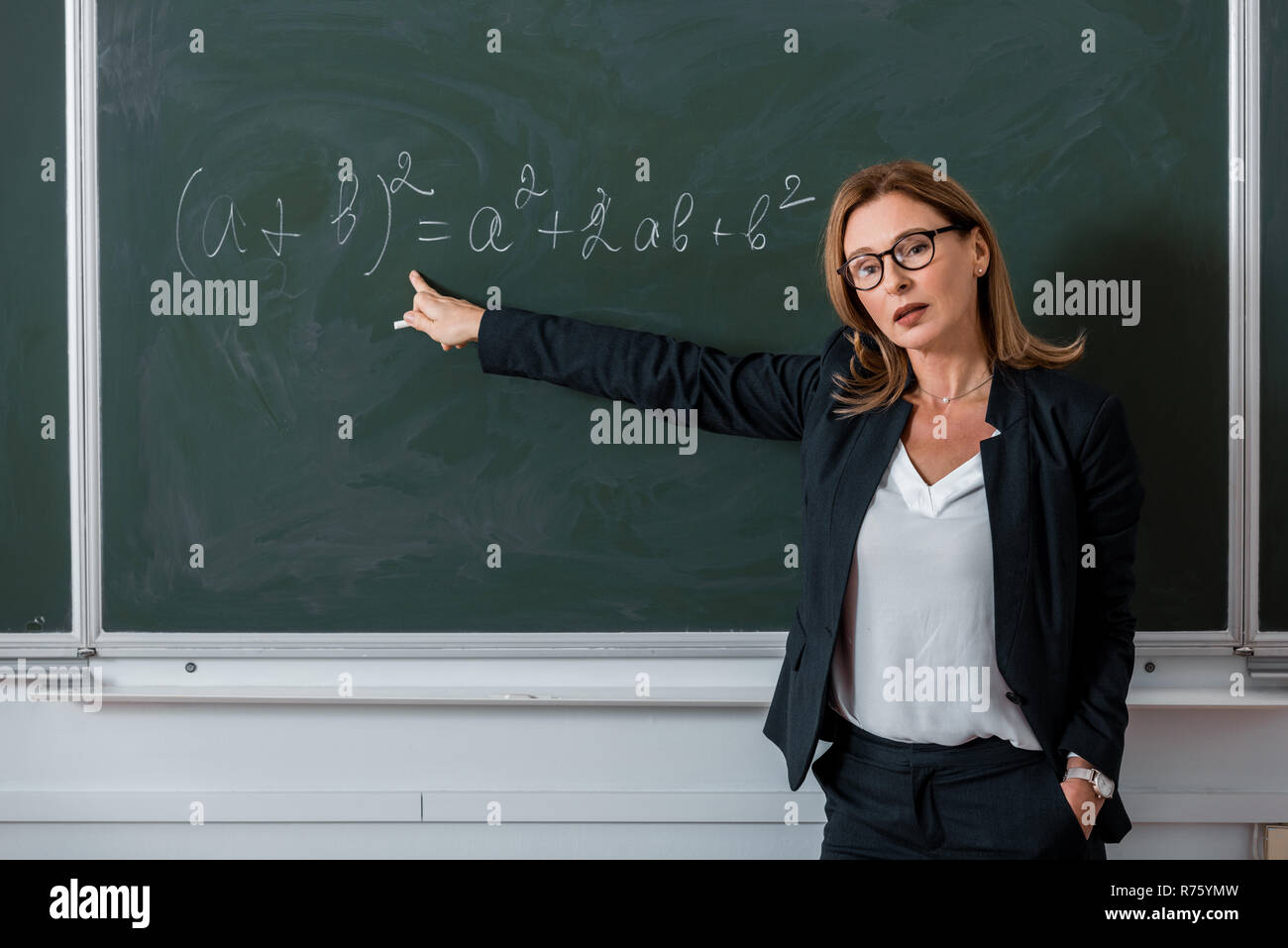 Insegnante femminile guardando la fotocamera e puntando con il dito in corrispondenza di equazione matematica sulla lavagna in classe Foto Stock