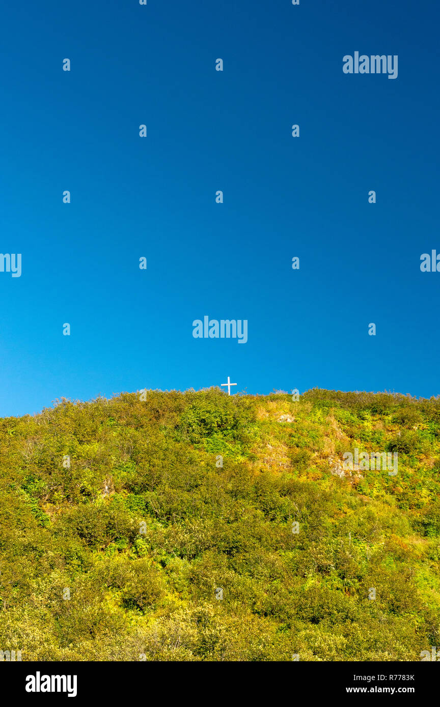 Piccola Croce Cristiana su una montagna verde in cima contro un vasto cielo blu chiaro. Foto Stock