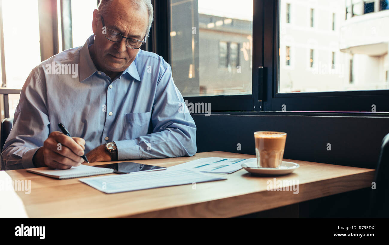 Imprenditore Senior udienza presso la caffetteria tabella e scrivere note sul blocco note. Uomo al lavoro da un moderno cafe'. Foto Stock