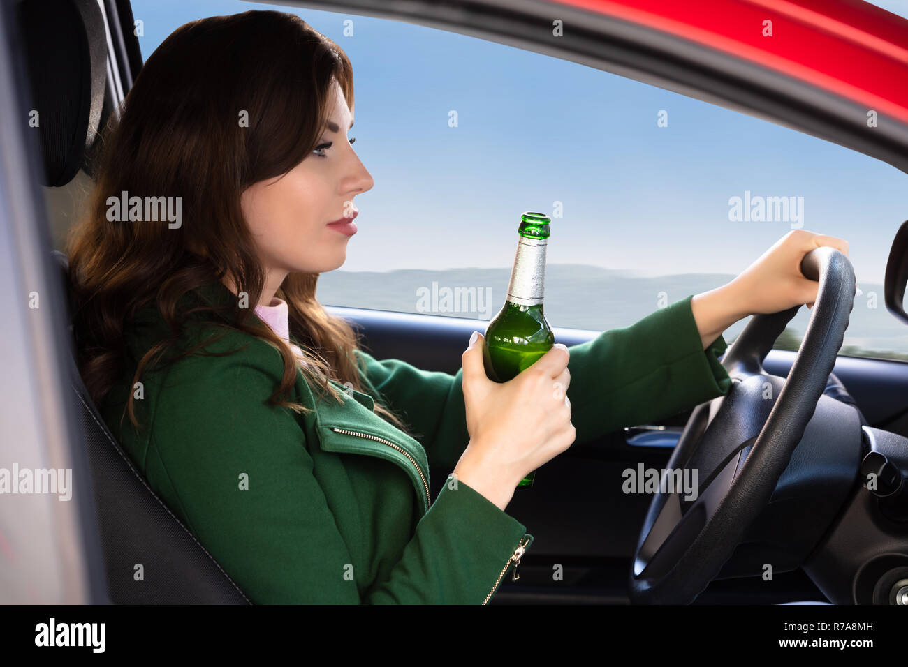 Vista laterale di una giovane e bella donna che mantiene la bottiglia di birra mentre si guida auto Foto Stock