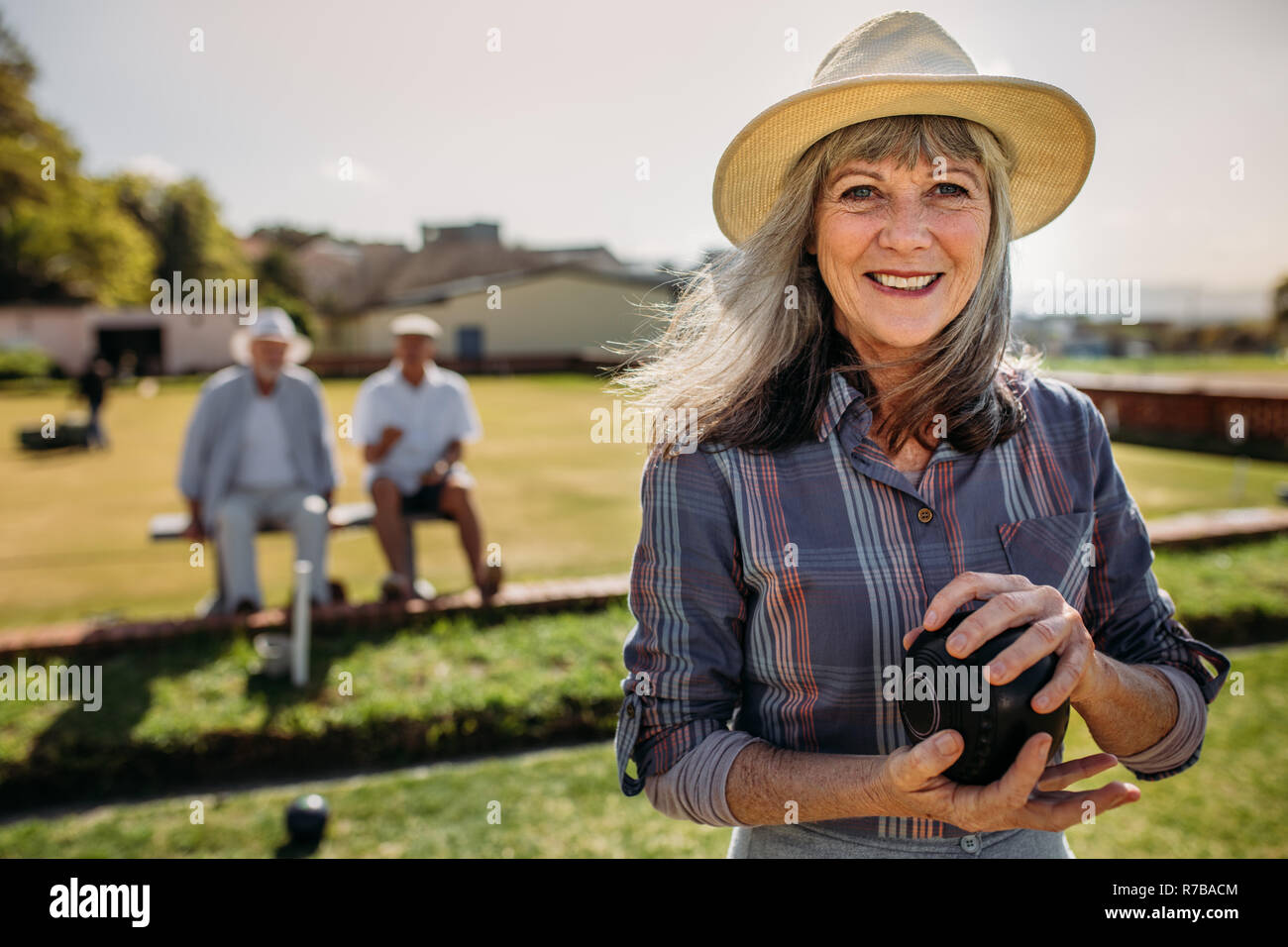 Sorridente donna senior in hat in piedi in un prato tenendo un bocce. Donna allegra giocando a bocce in un parco mentre i suoi amici maschi sono seduti in bac Foto Stock