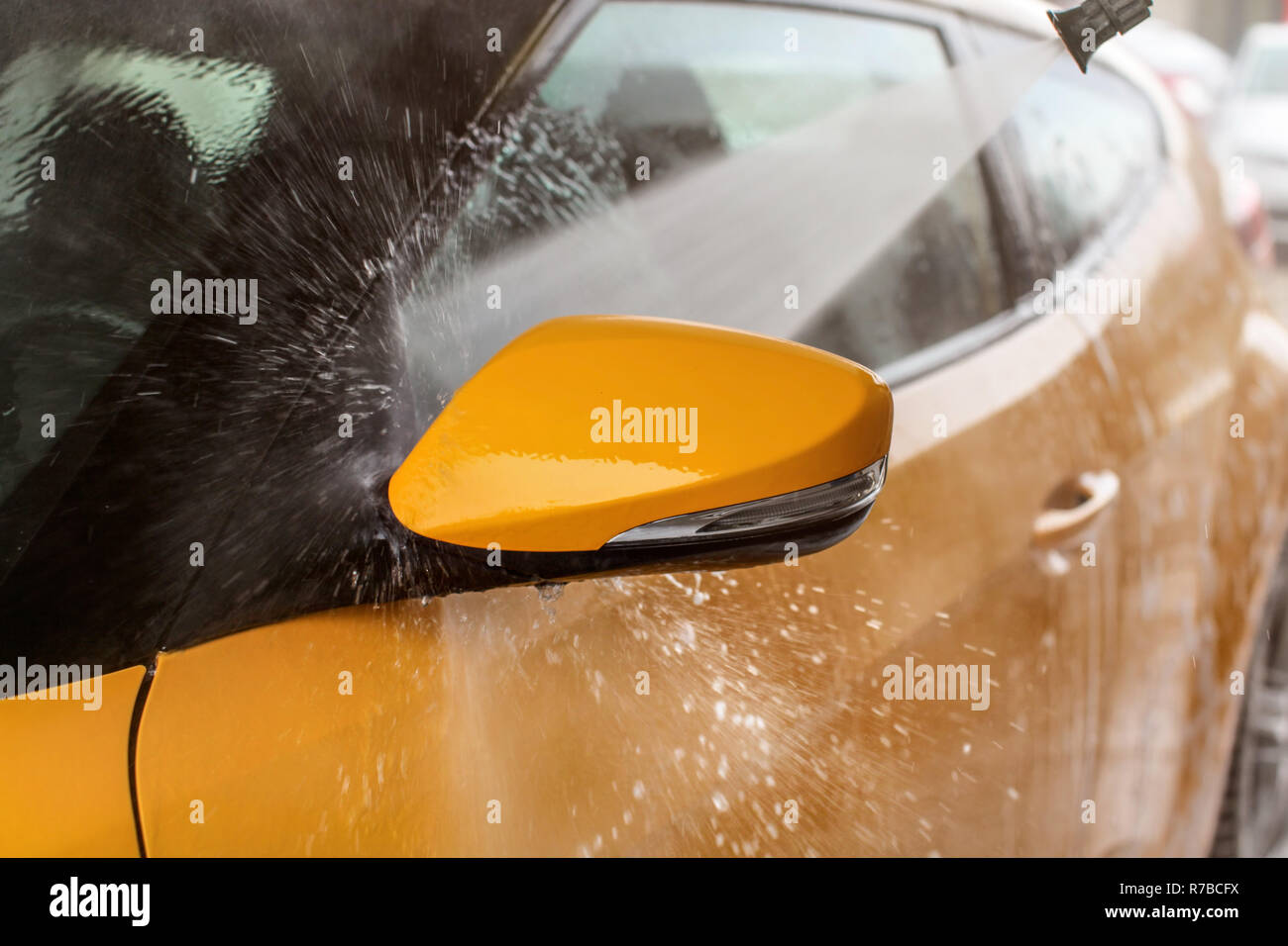 Lo specchietto laterale di giallo auto lavata in self service autolavaggi, spruzzo a getto di acqua che colpisce la finestra di gocce di acqua volare da tutti i lati. Foto Stock