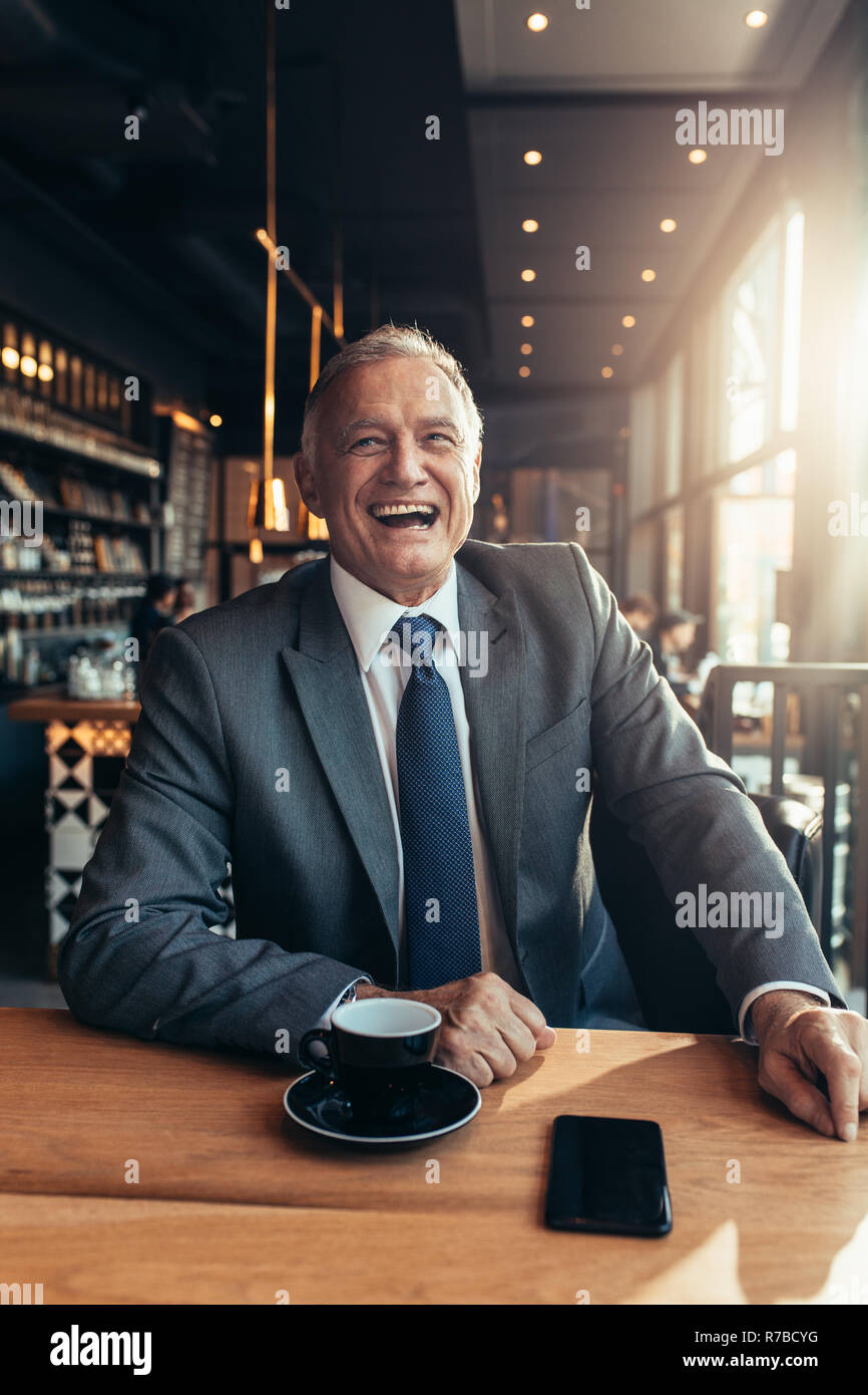 Ritratto di Allegro senior uomo in tuta seduti ad un ristorante con tazza di caffè e smart phone sul tavolo. Ridendo imprenditore presso la caffetteria. Foto Stock