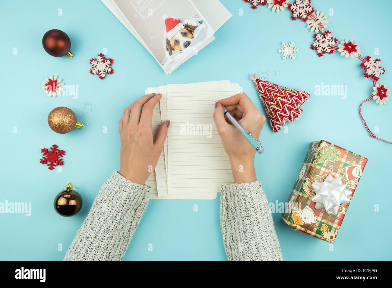 La scrittura di Natale lettera di saluto. Vista dall'alto di mani femminili con penna su sfondo blu con carta, holiday puntelli e doni e nuovo anno card Foto Stock