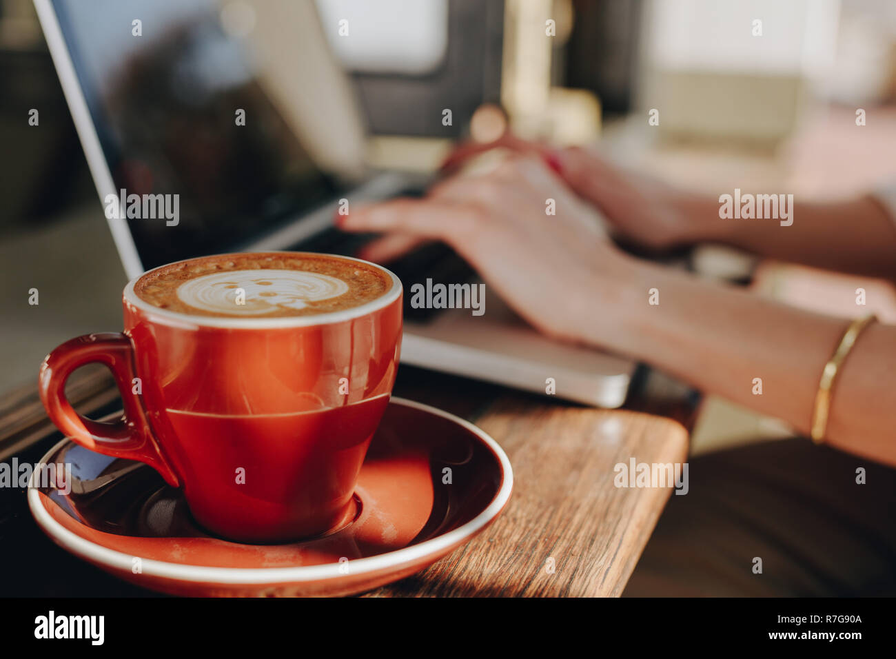 Tazza di caffè sul tavolo con donna seduta lavorando sul computer portatile. Donna che utilizza il computer portatile presso il cafe con tazza di caffè di fronte. Foto Stock