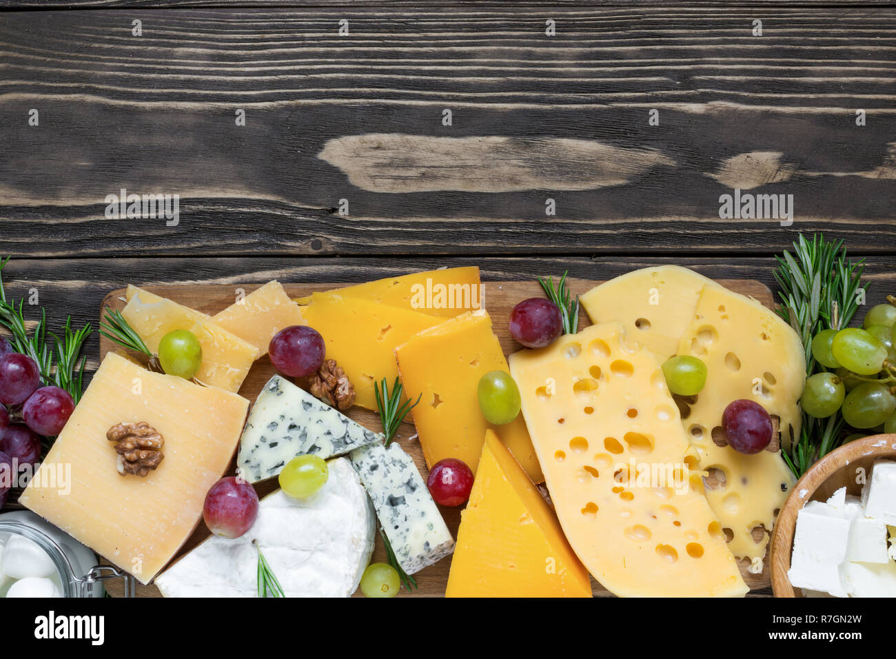 Vari tipi di formaggi sul tagliere di legno su tavola in legno rustico con uve e rosmarino, vista dall'alto Foto Stock