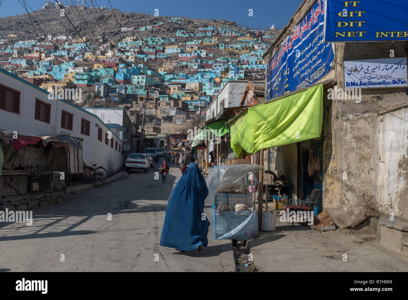 Signora indossando Burqa blu a camminare su una strada che conduce al dipinta di fresco colorate case di collina, Kabul, provincia di Kabul, Afghanistan Foto Stock