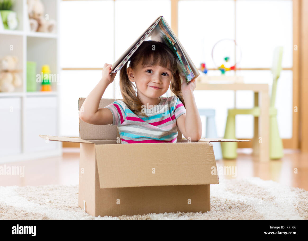 Smart kid ragazza seduta in una scatola di cartone e in possesso di un libro sopra la testa come tetto Foto Stock