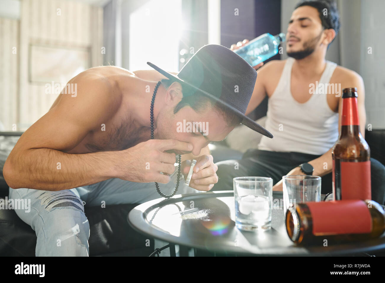 Tossicodipendente uomo sniffare la cocaina con la banconota arrotolata al partito a casa con un altro uomo di bere vodka in background Foto Stock