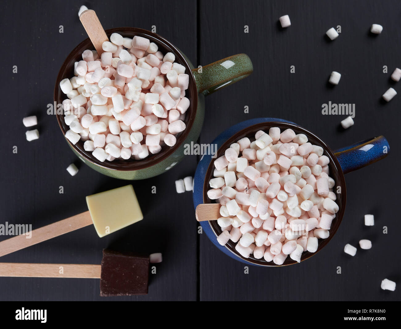 Due tazze con cioccolata calda e mini marshmallows, una tazza essendo blu e l'altra verde con il bianco e il cioccolato fondente a cubetti, sul dorso bruno scuro Foto Stock