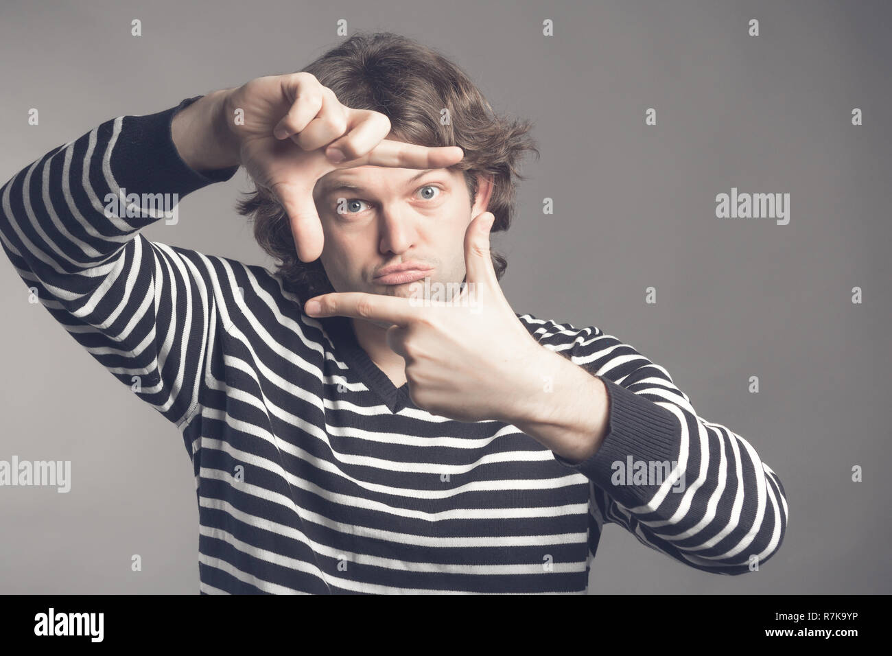 Giovane bello shaggy curly uomo la messa a fuoco con le dita, inquadratura con le mani su uno sfondo grigio, copia dello spazio. Close up ritratto uomo in maglione a strisce tiene le mani in un gesto di framing. Foto Stock