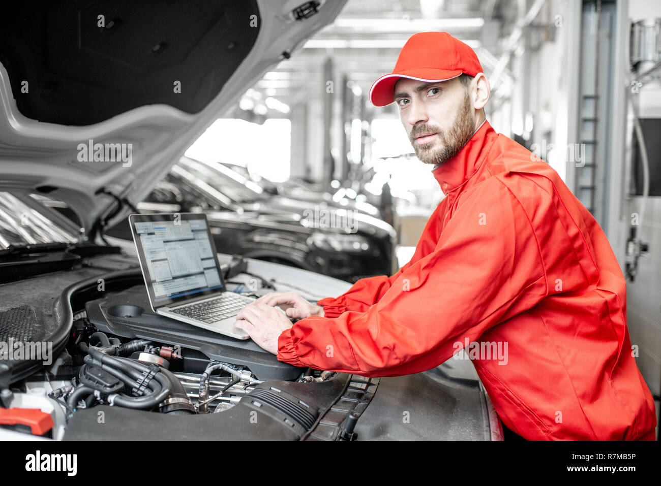 Ritratto di un bel meccanico automatico in rosso uniforme facendo la diagnostica motore con il calcolatore in auto di servizio Foto Stock