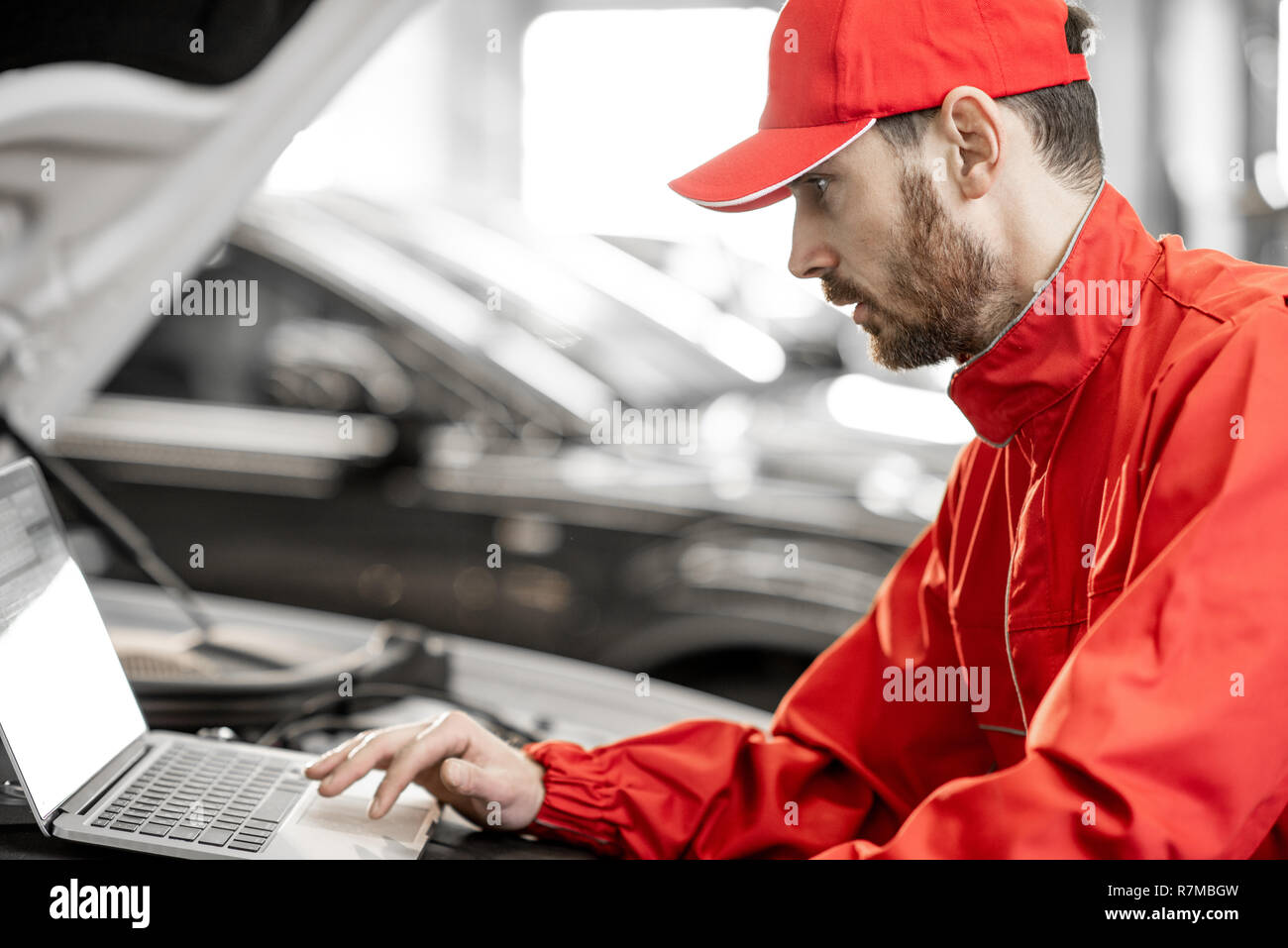 Bello meccanico automatico in rosso uniforme facendo la diagnostica motore con il calcolatore in auto di servizio Foto Stock