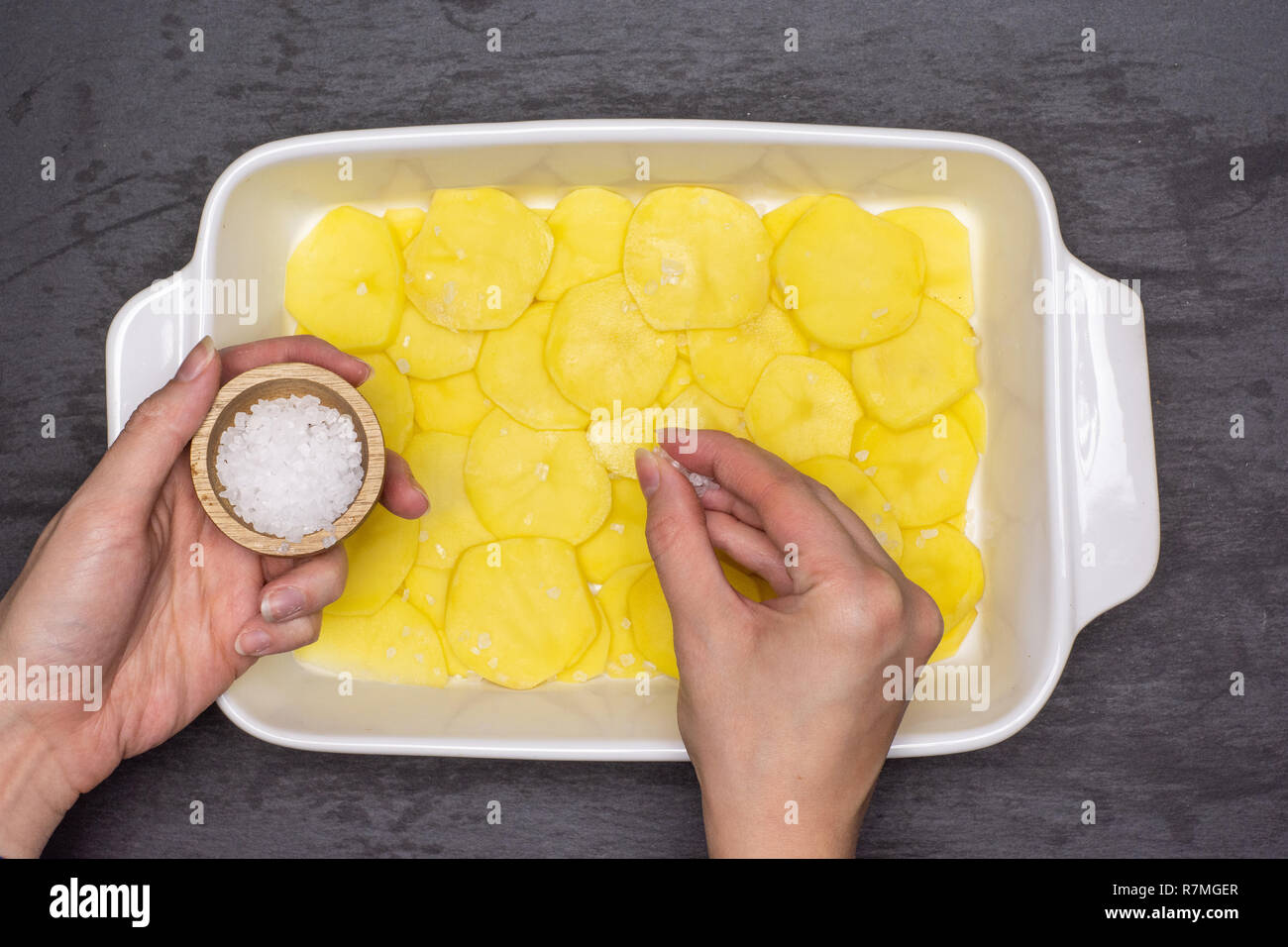 Donna mani sono strati di salatura. ricetta passo dopo passo le patate al forno con cipolla in ceramica bianca tostatore flatlay sulla pietra grigia Foto Stock