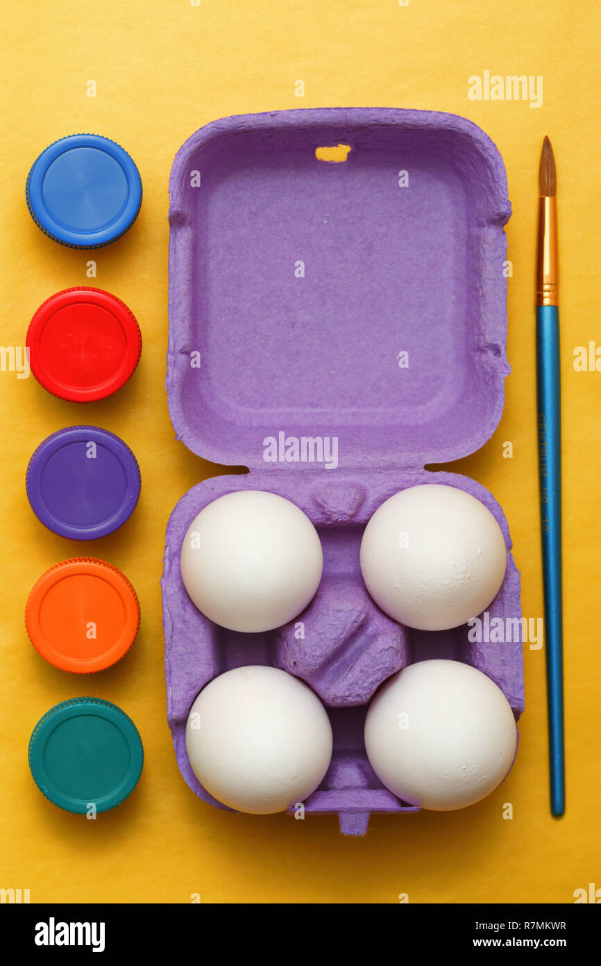 Composizione di pasqua. Set di quattro uova di colore bianco in una casella di colore viola, multi-colore di barattoli di vernice e un pennello per disegnare su sfondo giallo Foto Stock