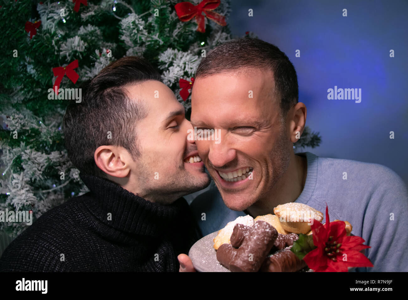 Maschio di coppia gay kissing, sorridente, ridendo di fronte ad albero di natale con la piastra di torte Foto Stock