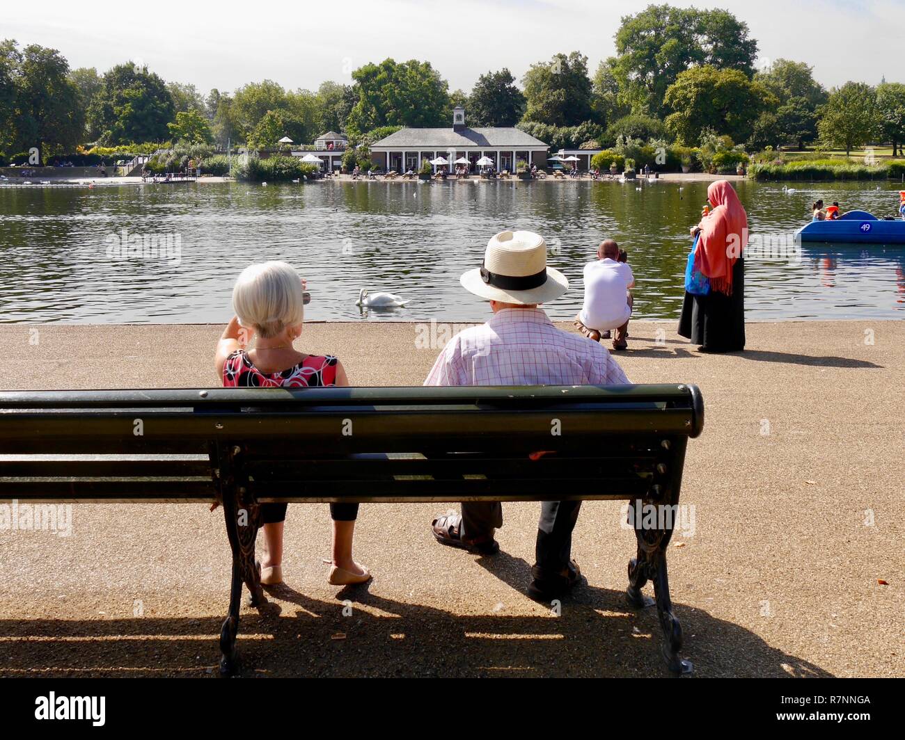 Coppia senior seduta sul banco da parte della serpentina, Hyde Park, Londra in estate con la donna in hijab in background in piedi al bordo d'acqua Foto Stock
