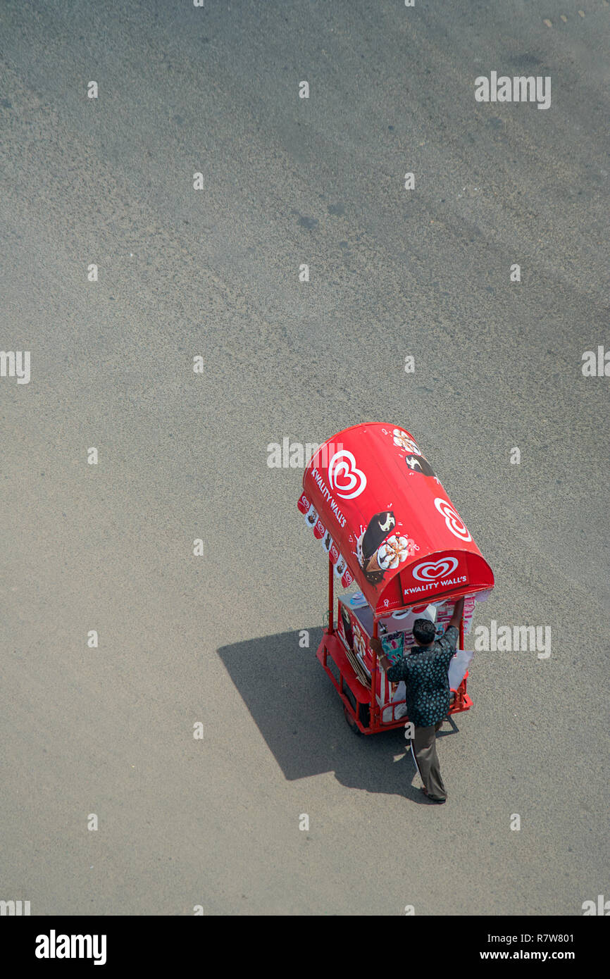 Di seguito su un asfalto street un maschio solitario street trader spinge il suo colore rosso brillante Gelato trolly lungo. Foto Stock