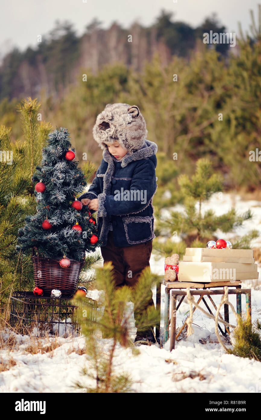 Un bambino decorare un albero di Natale all'aperto. Il ragazzo si blocca red bolle e mangia gingerbreads. Egli è vestito con un cappotto di pelle di pecora, nel verde dello sfondo Foto Stock