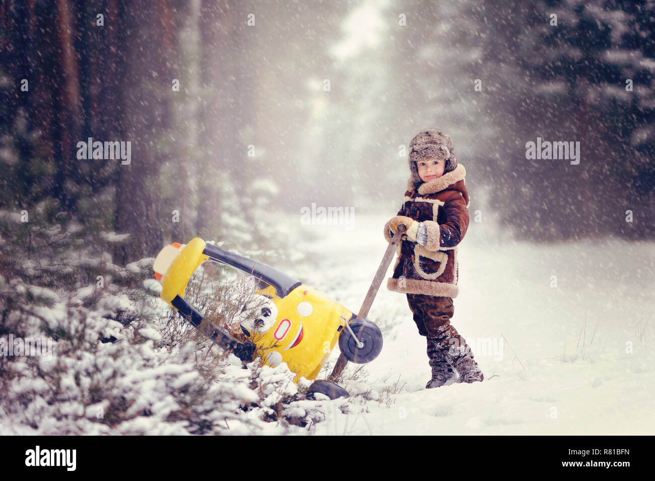 Bambino che gioca all'aperto durante la nevicata. Il ragazzo spinge dalla neve derive poco giallo auto, con due orsacchiotti interno, splendido nevoso inverno par Foto Stock