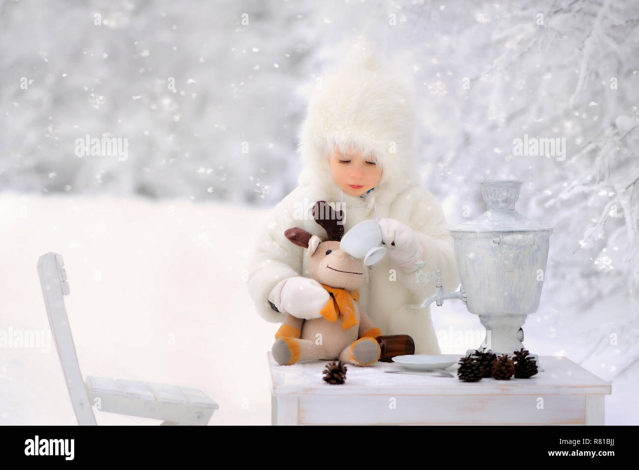 Il bambino in un bianco di rivestimento di pelle di pecora e cappuccio, apre un regalo e decora l'albero di Natale in una coperta di neve in inverno foresta. Un albero di Natale si erge Foto Stock