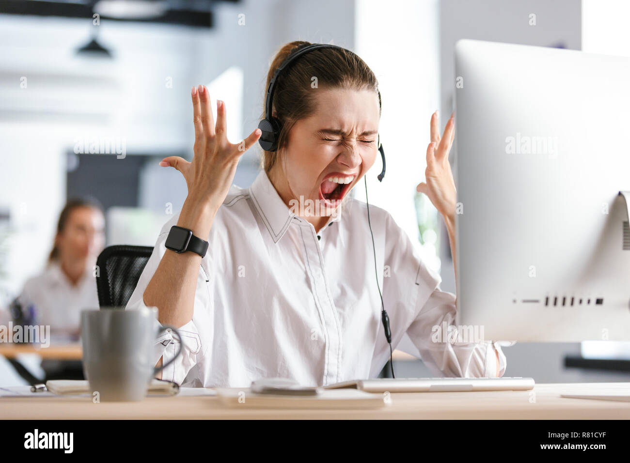 Arrabbiato giovane donna indossa cuffie con microfono vestito in camicia seduto al suo posto di lavoro presso l'ufficio, urlando Foto Stock