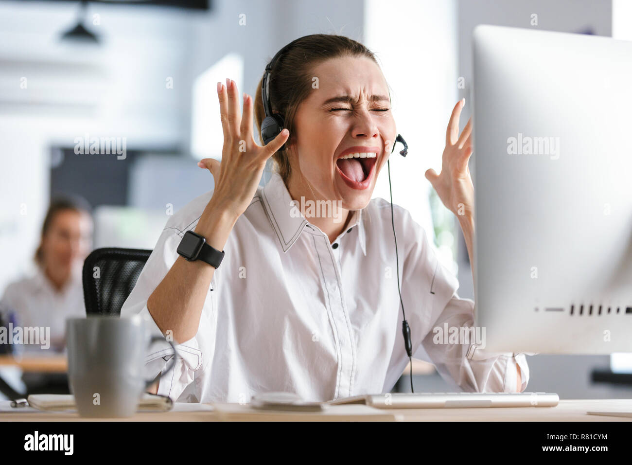 Arrabbiato giovane donna indossa cuffie con microfono vestito in camicia seduto al suo posto di lavoro presso l'ufficio, urlando Foto Stock