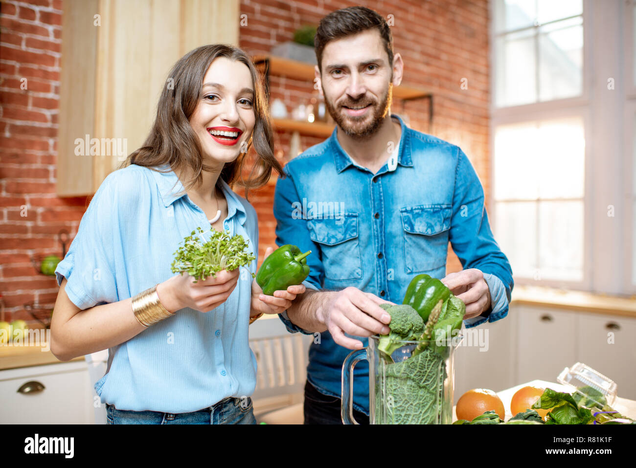 Ritratto di una giovane coppia di vegetariani in magliette blu rendendo smoothie con freschi prodotti verde sulla cucina a casa Foto Stock