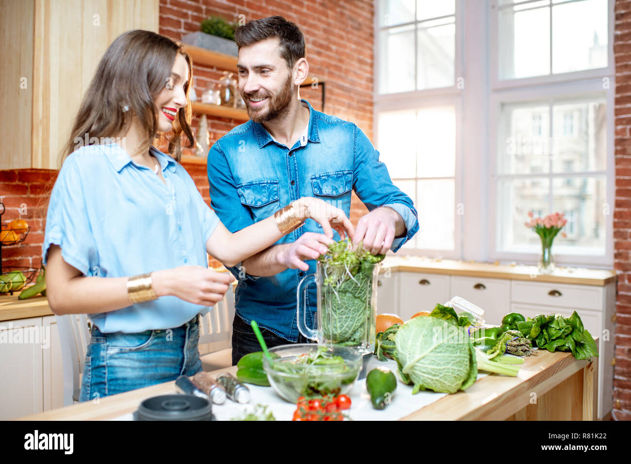 Coppia giovane di vegetariani in magliette blu rendendo smoothie con freschi prodotti verde sulla cucina a casa Foto Stock