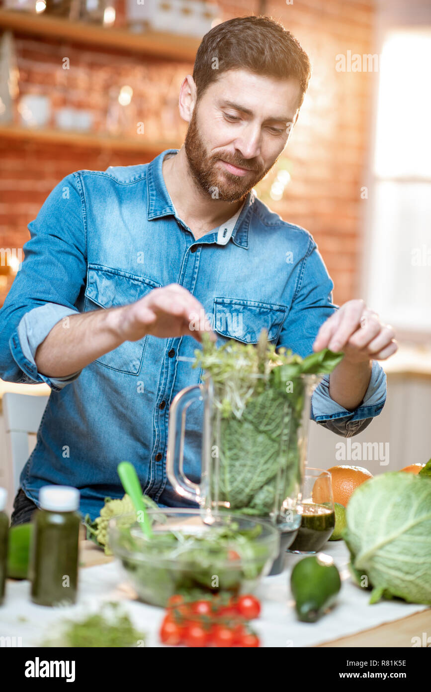 Uomo bello il riempimento di Blender con verde cibo crudo, rendendo frullato vegetariano sulla cucina a casa Foto Stock