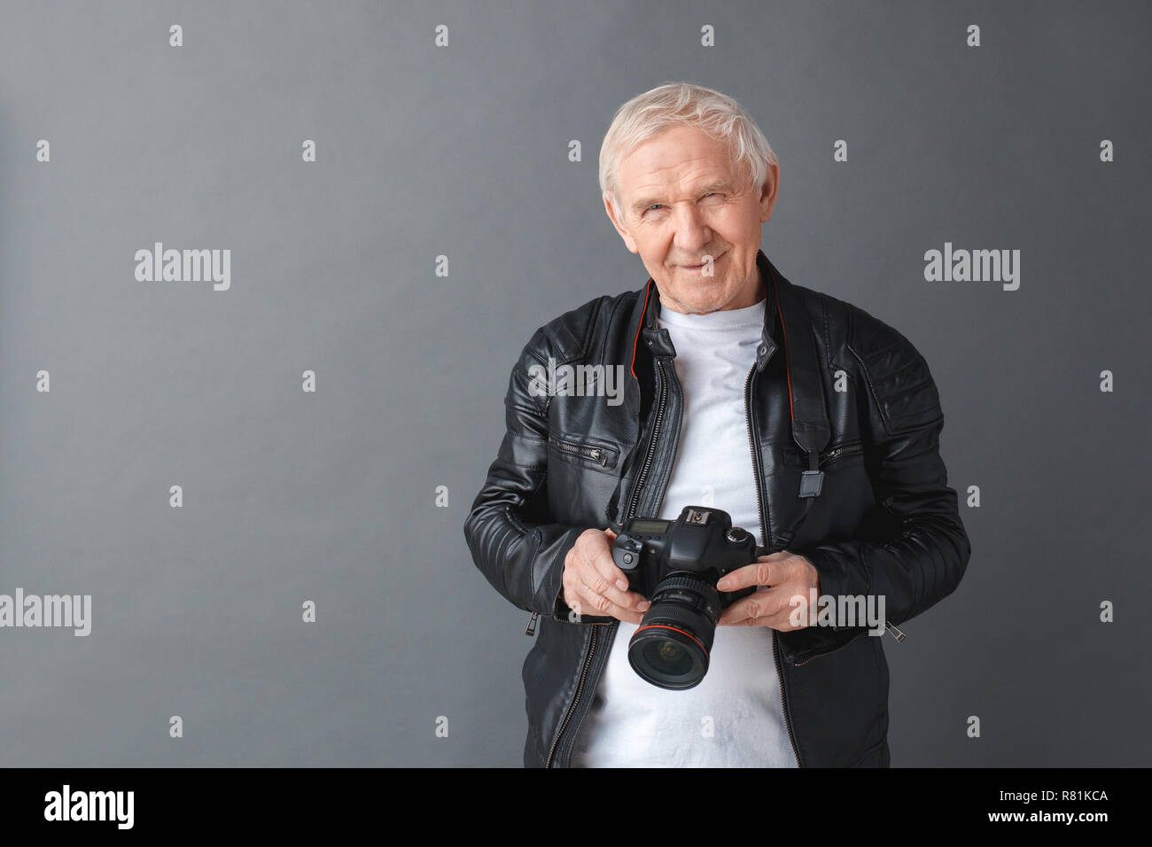 Senior uomo in giacca di pelle in piedi isolato su grigio tenere la fotocamera giocoso Foto Stock