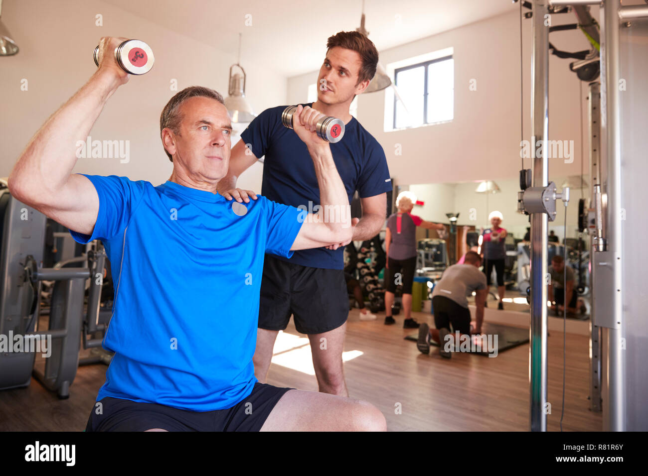 Senior uomo esercizio con pesi essendo incoraggiati da Personal Trainer in palestra Foto Stock