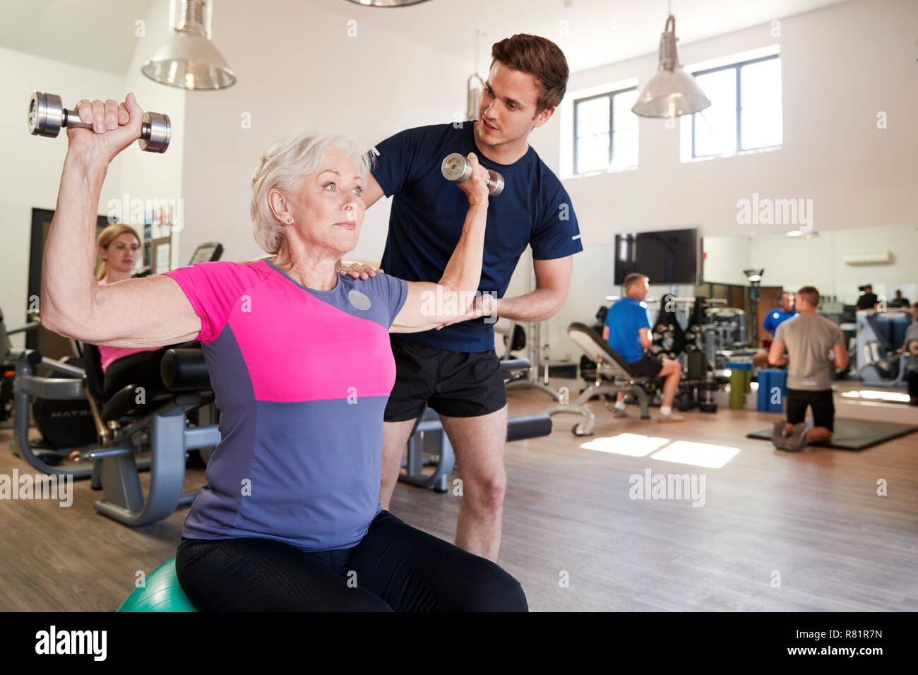 Senior donna esercita sulla sfera svizzero con pesi essendo incoraggiati da Personal Trainer in palestra Foto Stock