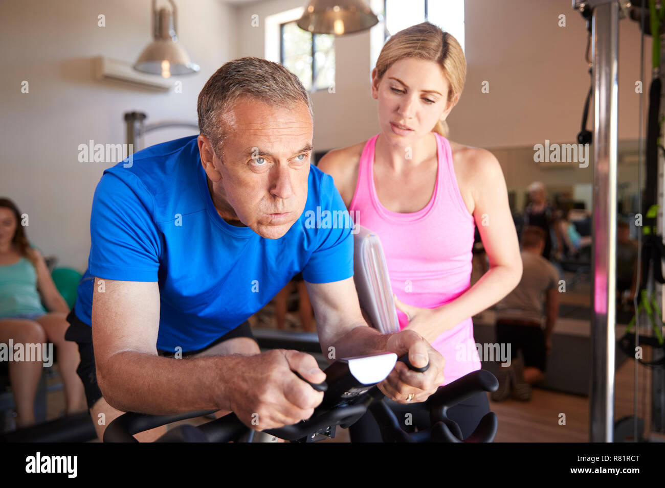 Senior uomo esercita sul ciclismo macchina essendo incoraggiati dalla femmina Personal Trainer in palestra Foto Stock