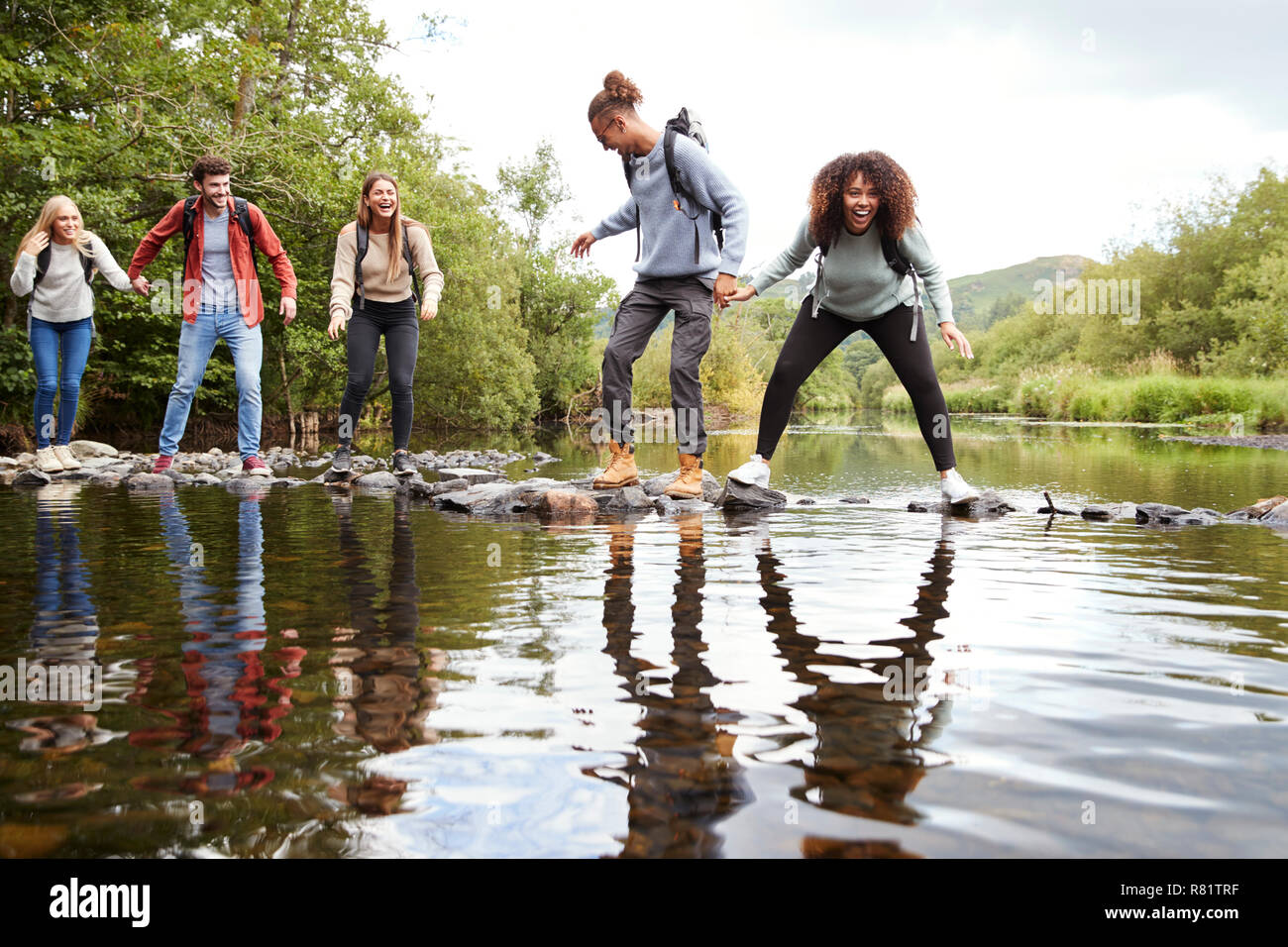 Multi etnico gruppo di cinque giovani amici adulti a ridere come loro equilibrio sulle rocce di attraversare un flusso durante una escursione Foto Stock