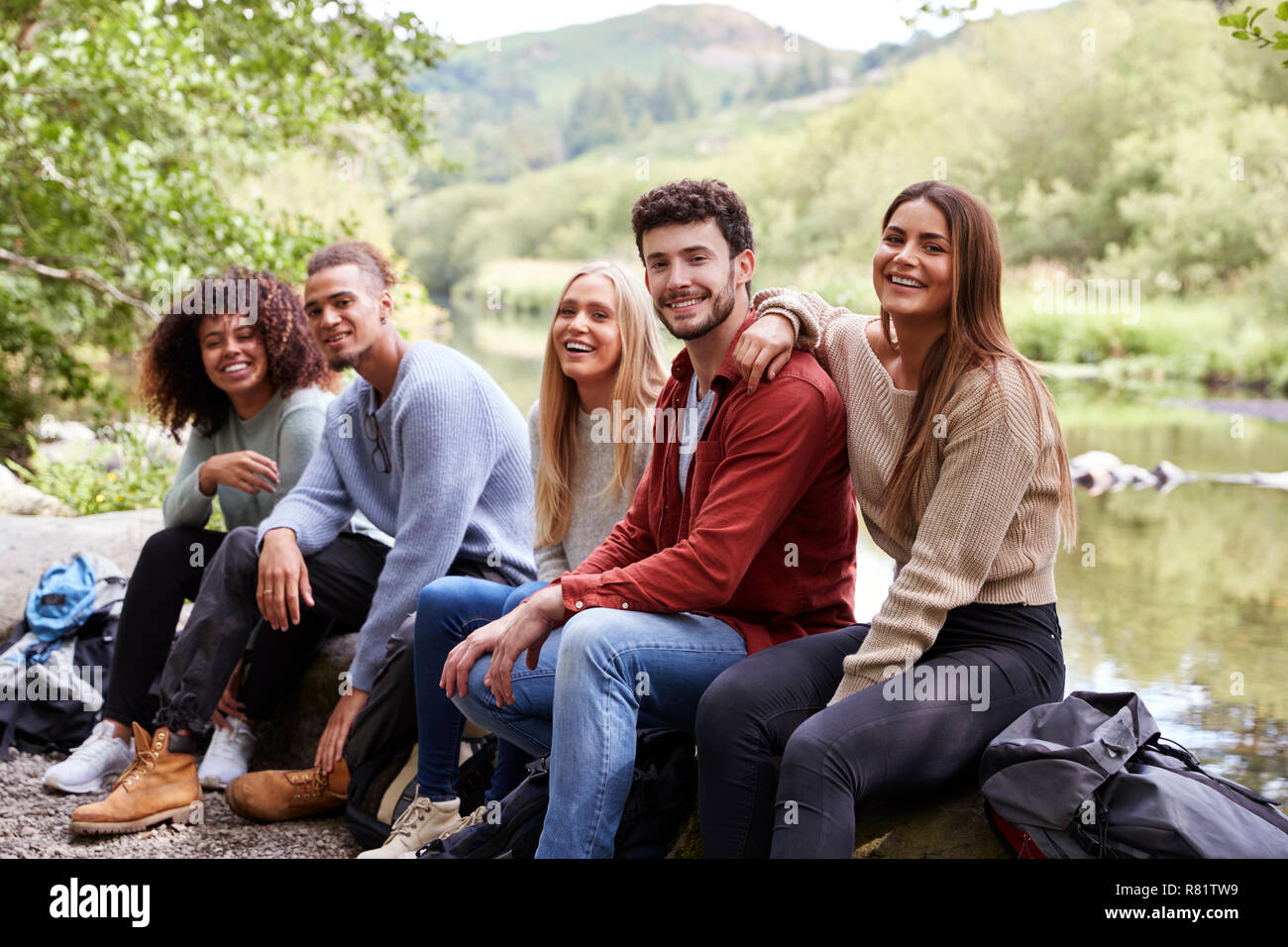 Multi etnico gruppo di cinque giovani amici adulti prendendo una pausa seduta sulle rocce da un flusso durante un'escursione, ritratto Foto Stock
