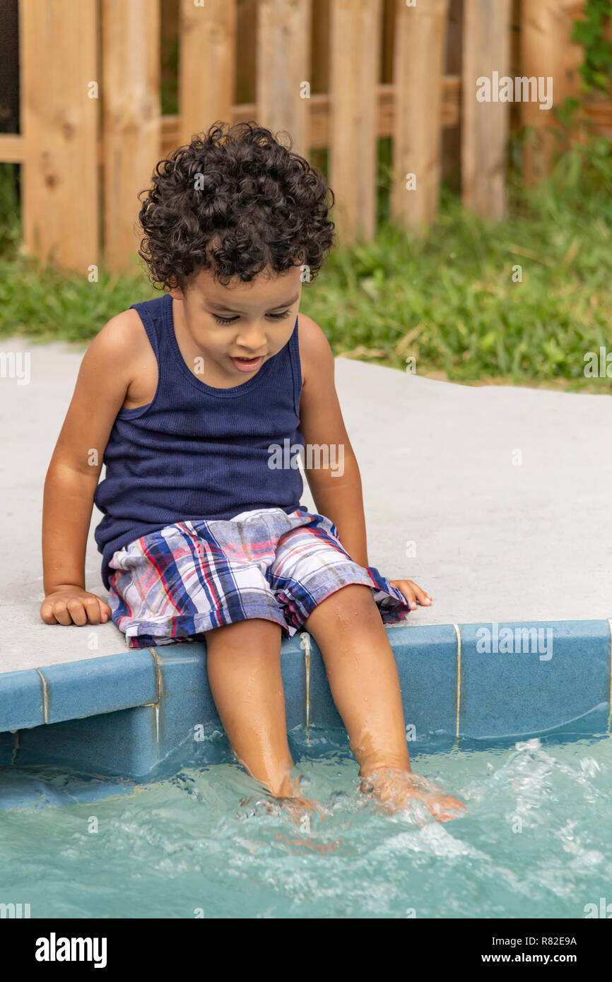 Un bimbo stupito imposta a bordo piscina con i piedi nella piscina. Un ragazzo ispanica è stupito a l'acqua in movimento a cascata sui suoi piedi. Foto Stock